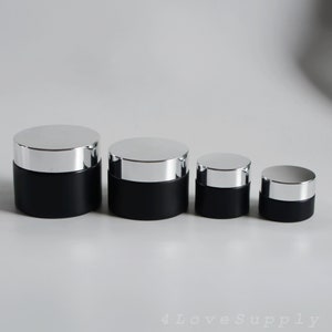 Op de afbeelding: Vier cilindervormige cosmetische potjes gerangschikt in afnemende grootte. Elk potje heeft een matzwarte basis en een reflecterend zilverkleurig deksel. De tekst 4 Love Supply is zichtbaar in de rechterbenedenhoek van de afbeelding in een licht lettertype.