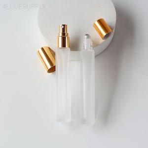 May include: Two clear, rectangular perfume bottles with gold-colored caps and spray nozzles. One bottle has a rollerball applicator. The bottles are on a white surface. The text "4LOVESUPPLY" is in the upper left corner.