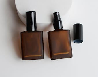May include: Two amber glass perfume bottles with black spray tops and a separate black cap. The bottles are rectangular and sit on a white surface. One bottle has a spray nozzle, and the other has a solid black top.