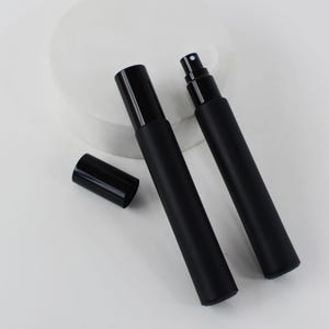 May include: Two black perfume atomisers with matching caps. One atomiser has its cap off, revealing the opening. The other atomiser has a spray nozzle. The atomisers are on a white surface.