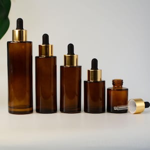 Könnte beinhalten: Eine Sammlung von Braunglas-Tropfflaschen in verschiedenen Größen. Jede Flasche hat einen goldfarbenen Kragen und eine schwarze Gummitropfer. Die Flaschen sind in einer Reihe angeordnet, um ihre unterschiedlichen Höhen und Kapazitäten zu zeigen. Der Hintergrund ist schlicht weiß.