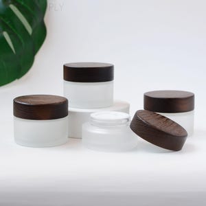 Op de afbeelding: Een verzameling glazen potten met een matte afwerking en donkerbruine houten deksels. Eén pot heeft het deksel eraf, waardoor de binnenkant zichtbaar is. De potten zijn van verschillende formaten en staan op een witte ondergrond.