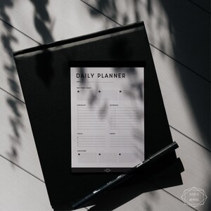 May include: A black and white daily planner with the text "DAILY PLANNER" at the top. The planner has sections for date, top three tasks, checklist, schedule, meals, notes, and gratitude.