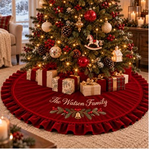 May include: A round, deep red velvet Christmas tree skirt with a ruffled edge. The skirt features embroidered green pine branches, red berries, and a gold bell, with the text The Watson Family stitched in elegant white script lettering.