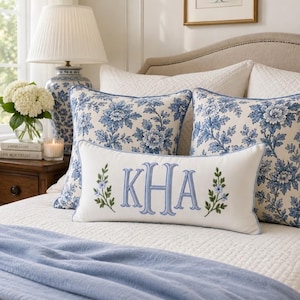 Puede incluir: Un cojín lumbar rectangular blanco con el monograma azul bordado KHA flanqueado por dos pequeñas ramas florales verdes y azules, colocado frente a dos cojines cuadrados con un patrón floral azul y blanco sobre una cama.