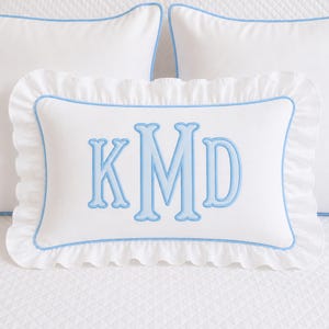 May include: A rectangular white throw pillow featuring a ruffled edge and light blue piping. The center of the pillow displays a large, embroidered monogram with the letters KMD in a light blue font, matching the piping and ruffle trim.