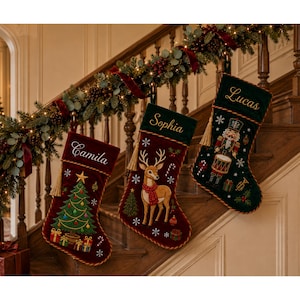 Könnte beinhalten: Drei Weihnachtssocken aus rotem und dunkelgrünem Samt mit goldener Verzierung und Quasten. Die Socken sind mit den Namen Camila, Sophia und Lucas personalisiert und zeigen gestickte Motive eines Weihnachtsbaums, eines Hirsches und eines Nussknackers.