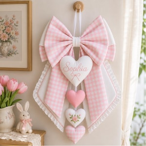 May include: A decorative wall hanging featuring a large pink and white gingham bow with white ruffled edges. Suspended from the centre are four plush hearts: the top one is white with the name Sophia printed in pink, followed by three smaller hearts.
