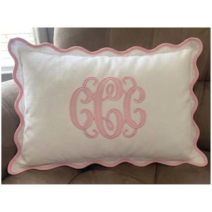 May include: A rectangular white throw pillow featuring a scalloped edge trimmed in light pink. The centre of the pillow displays a large, embroidered monogram consisting of three interlocking letters in a matching light pink thread colour.