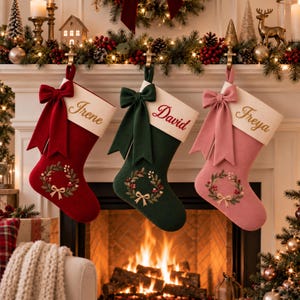 Puede incluir: Tres calcetines navideños en rojo, verde oscuro y rosa empolvado, cada uno con un puño color crema bordado con los nombres Irene, David y Freya. Cada calcetín está adornado con un gran lazo a juego y un bordado de corona floral.