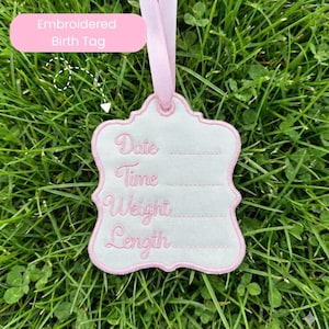 May include: Embroidered birth tag with a pink ribbon, featuring a white background and pink trim. The tag includes embroidered text fields for date, time, weight, and length. The tag is displayed on a bed of green grass.