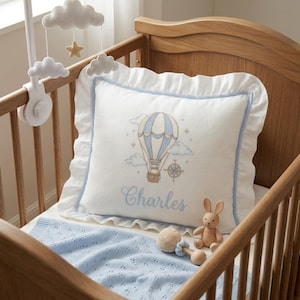 May include: A white baby pillow with a blue and white hot air balloon design, the name "Charles" embroidered in blue, and a blue ruffle trim. The pillow is on a blue blanket in a wooden crib, with a mobile and stuffed animals.