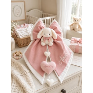 Può includere: Un fiocco in tessuto rosa e bianco con un coniglietto di peluche color crema attaccato al centro. Il fiocco ha un bordo bianco ed è ricamato con il nome Amelia. Due ornamenti a forma di cuore pendono dal coniglio, uno ricamato Amelia.