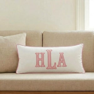 Op de afbeelding: Een rechthoekig wit sierkussen met de geborduurde roze monogramletters HLA in het midden. Het kussen is rondom afgewerkt met een bijpassende roze bies, wat zorgt voor een strakke en duidelijk gedefinieerde rand aan het ontwerp.