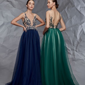 May include: Two floor-length sleeveless gowns featuring sheer illusion bodices with dark floral embroidery. One gown is deep navy blue with a plunging neckline, while the other is emerald green with a low back, both with full tulle skirts.