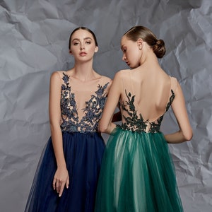 May include: Two sleeveless floor-length gowns featuring sheer illusion bodices with dark floral embroidery. One gown is navy blue with a deep V-neckline, while the other is emerald green with a low-cut back and a full, layered tulle skirt.