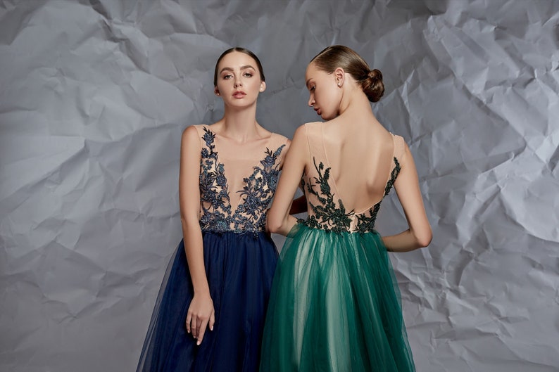 May include: Two sleeveless floor-length gowns featuring sheer illusion bodices with dark floral embroidery. One gown is navy blue with a deep V-neckline, while the other is emerald green with a low-cut back and a full, layered tulle skirt.