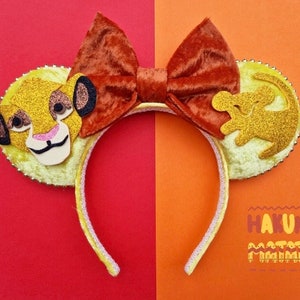May include: Yellow mouse ears headband with a brown velvet bow. One ear features a cartoon image of Simba from The Lion King, the other ear features a cartoon image of Simba as a cub. The headband has the text "Hakuna Matata" printed on it.