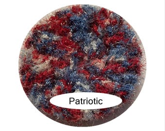 May include: A circular patch of textured, fuzzy yarn featuring a variegated blend of deep red, navy blue, and white fibers. The material has a shaggy, multi-dimensional appearance with a white oval label at the bottom displaying the text Patriotic.