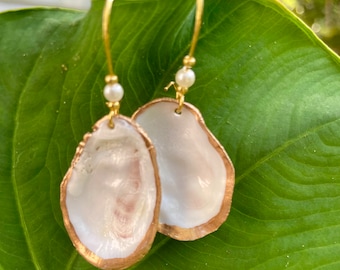 May include: Handmade earrings featuring oyster shells with gold-painted edges and gold-coloured ear wires. Each earring has a small, white pearl bead. The shells have a pearlescent white interior.