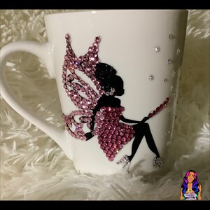 May include: White ceramic mug with a pink and black fairy design. The fairy has wings made of pink rhinestones and is sitting on a branch. The mug is decorated with small, clear rhinestones.