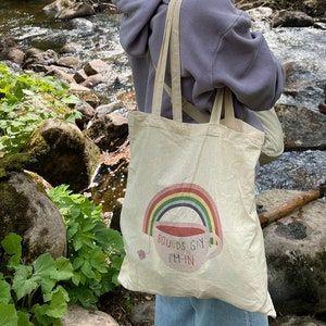 Könnte beinhalten: Beige Tragetasche mit Regenbogen-Design und dem Text "Sounds Gay I'm In". Die Tasche hat lange Träger und wird im Freien getragen. Der Regenbogen befindet sich über einer Teetasse mit Teebeutel.
