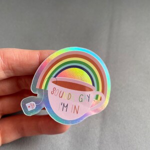 May include: Holographic sticker featuring a rainbow and a teacup with the text "SOUNDS GAY I'M IN". The teacup has a green shamrock on the side. A small tea bag with a heart is also visible.