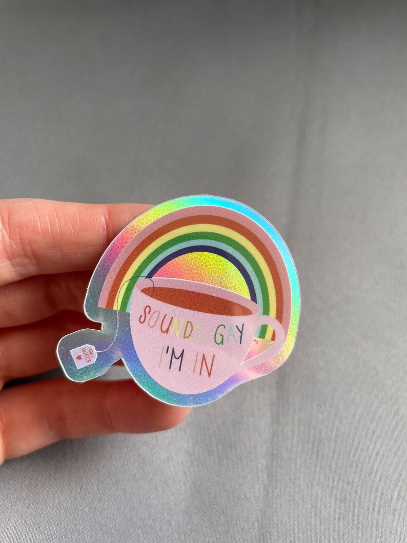 May include: Holographic sticker featuring a rainbow and a teacup with the text "SOUNDS GAY I'M IN". The teacup has a green shamrock on the side. A small tea bag with a heart is also visible.