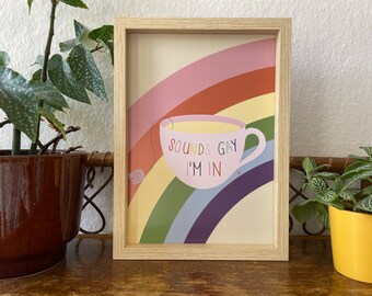 May include: A framed art print featuring a rainbow design with a teacup illustration. The teacup is pink and contains tea, with the text "SOUNDS GAY I'M IN". The background is a rainbow, and the frame is light wood.