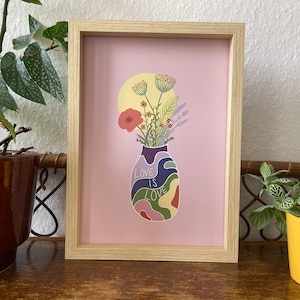 Może przedstawiać: Oprawiony wydruk artystyczny z wazonem kwiatów na różowym tle. Na wazonie napis "LOVE IS LOVE". Grafika w jasnej drewnianej ramie, odpowiednia do dekoracji wnętrz. Kolorowa ilustracja.