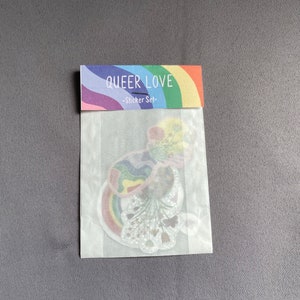 May include: A sticker set in a clear plastic package. The packaging has a rainbow design with the words "QUEER LOVE" and "-Sticker Set-". The stickers inside feature rainbow designs and floral elements.