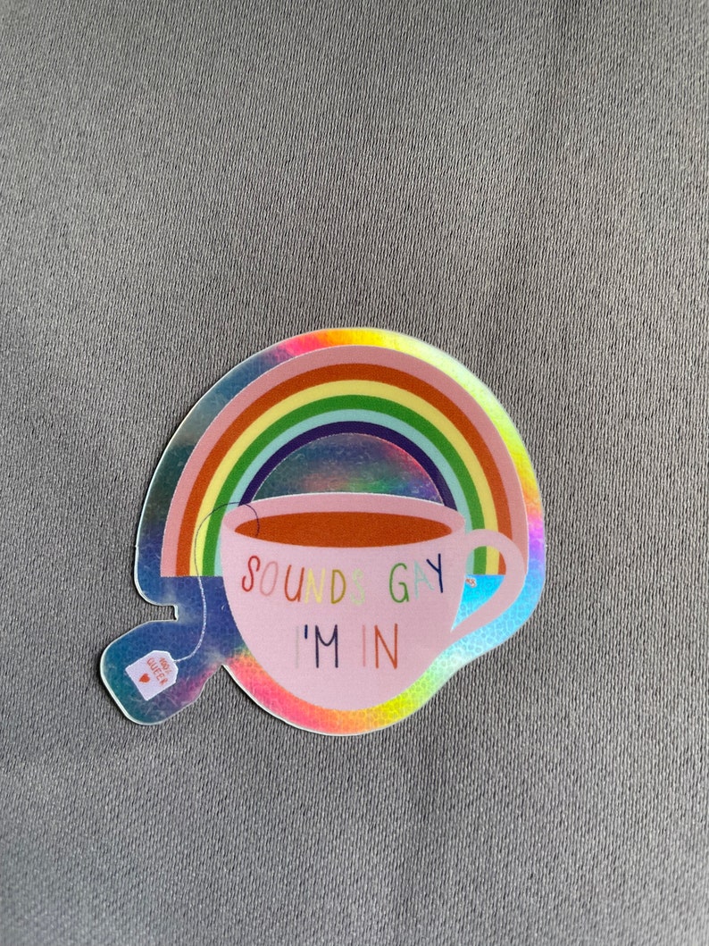 May include: A holographic sticker featuring a pink teacup with the text "SOUNDS GAY I'M IN" in colorful letters. A rainbow arches over the teacup. A small tea bag with the text "TEA QUEER" is attached to the cup.