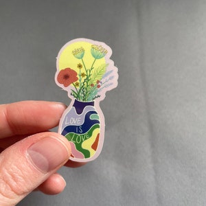 May include: A sticker featuring a vase-shaped design with a floral arrangement and the text "LOVE IS LOVE." The vase is filled with colorful flowers and foliage, with a yellow background. The sticker has a pink outline.