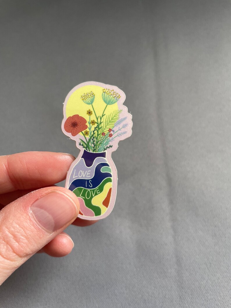 May include: A sticker featuring a vase-shaped design with a floral arrangement and the text "LOVE IS LOVE." The vase is filled with colorful flowers and foliage, with a yellow background. The sticker has a pink outline.