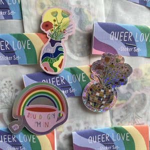 May include: A collection of colorful stickers with a queer theme. The stickers feature a rainbow, a teacup with the text "SOUND GAY I'M IN", and vases with floral arrangements. The stickers are packaged with "QUEER LOVE - Sticker Set-" cards.