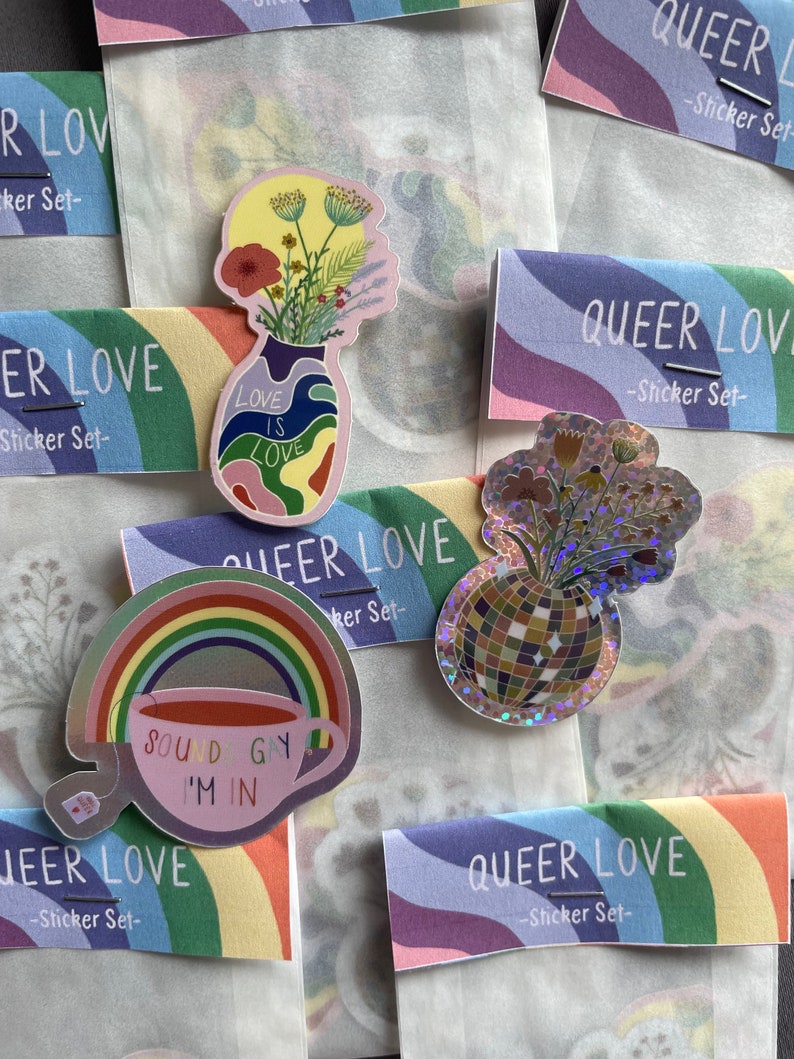 May include: A collection of colorful stickers with a queer theme. The stickers feature a rainbow, a teacup with the text "SOUND GAY I'M IN", and vases with floral arrangements. The stickers are packaged with "QUEER LOVE - Sticker Set-" cards.