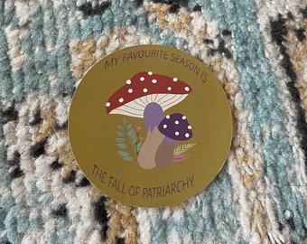 May include: A circular sticker with a mustard yellow background featuring two mushrooms, one large red with white spots and one smaller purple with white spots, surrounded by small green leaves. The text reads: MY FAVOURITE SEASON IS THE FALL OF PATRIARCHY.