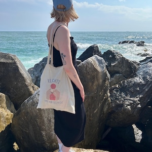 May include: A canvas tote bag with the German text "RECHTS" and an illustration of a person knitting. The bag is carried by a person wearing a black dress and a blue cap, standing on rocks near the ocean.