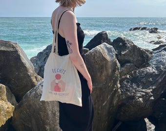 May include: A canvas tote bag with the German text "RECHTS" and an illustration of a person knitting. The bag is carried by a person wearing a black dress and a blue cap, standing on rocks near the ocean.