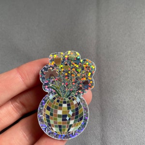 May include: A holographic sticker featuring a disco ball vase filled with dried flowers. The sticker has a glittery, iridescent border and a cloud-like shape around the flowers. The disco ball is made of squares in shades of green, brown, and blue.