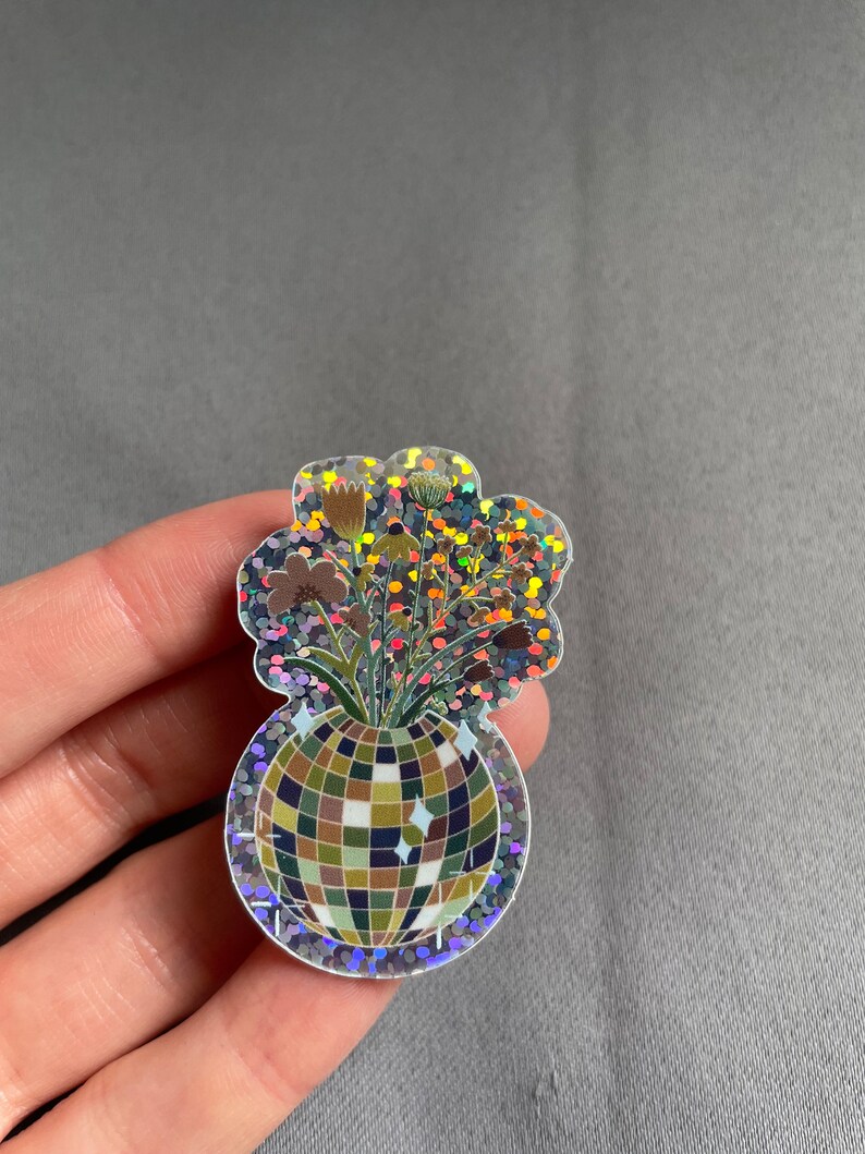 May include: A holographic sticker featuring a disco ball vase filled with dried flowers. The sticker has a glittery, iridescent border and a cloud-like shape around the flowers. The disco ball is made of squares in shades of green, brown, and blue.