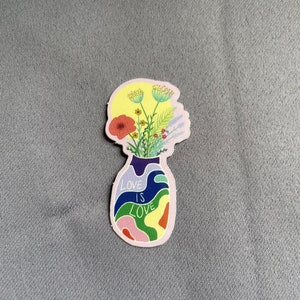 May include: A sticker featuring a vase-shaped design with the words "LOVE IS LOVE" in white. The vase is filled with colorful, abstract shapes and flowers. The flowers include a red poppy, yellow and green sprigs, and other colorful blooms.