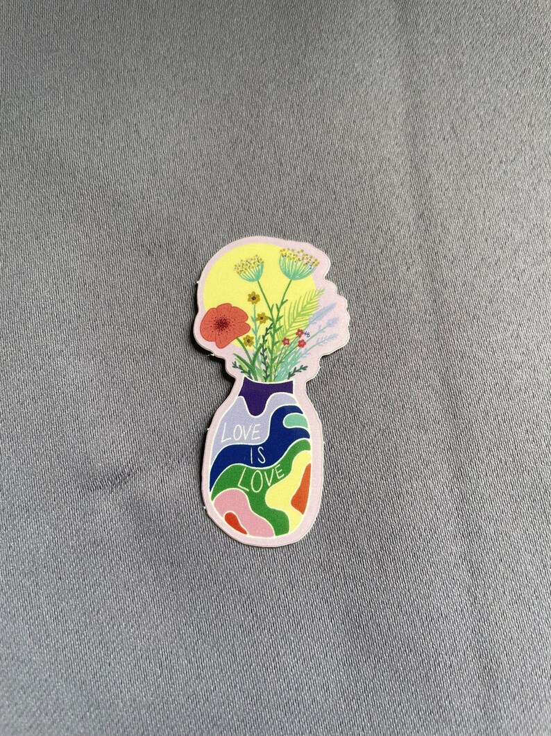 May include: A sticker featuring a vase-shaped design with the words "LOVE IS LOVE" in white. The vase is filled with colorful, abstract shapes and flowers. The flowers include a red poppy, yellow and green sprigs, and other colorful blooms.