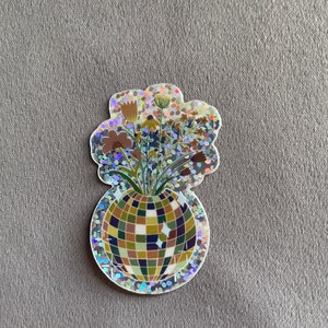 May include: A holographic sticker featuring a disco ball vase filled with flowers. The vase is a mosaic of green, brown, and blue squares. The flowers are brown, yellow, and green. The sticker has a glittery, iridescent border.