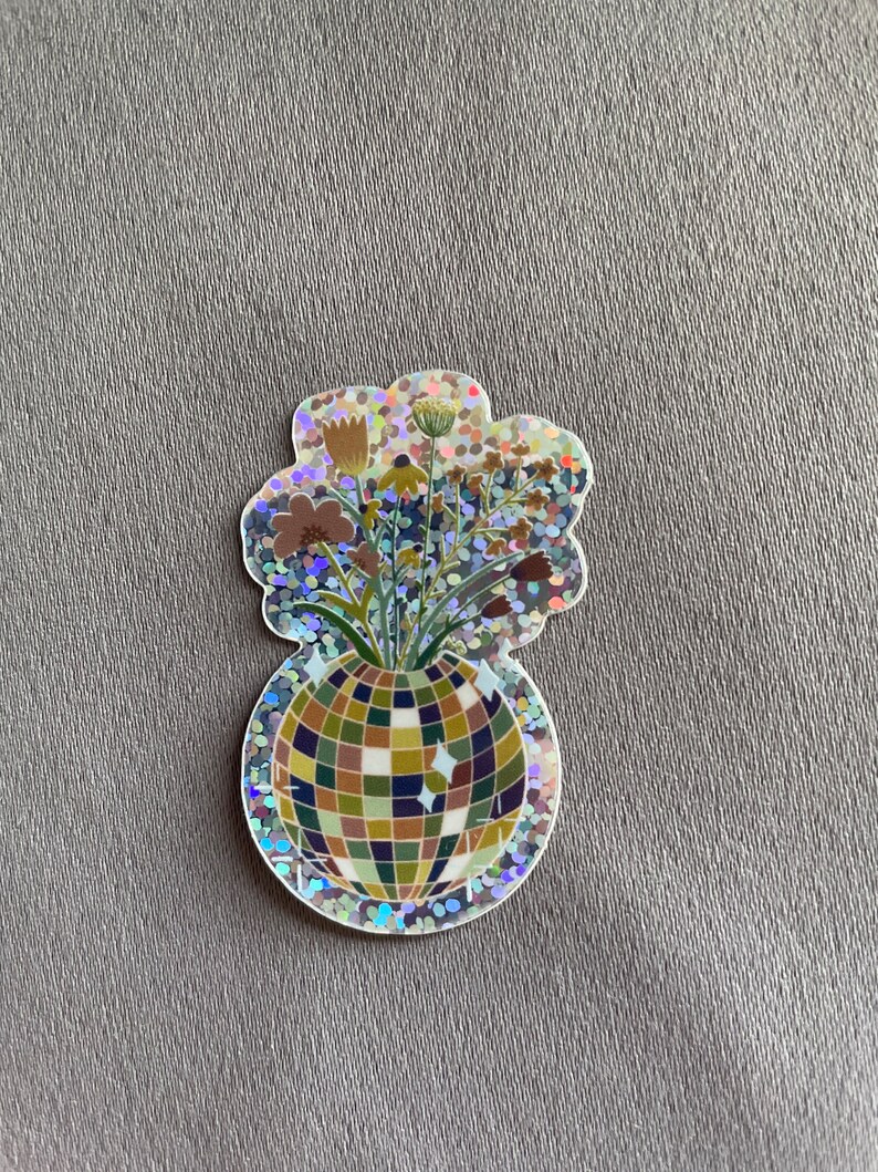 May include: A holographic sticker featuring a disco ball vase filled with flowers. The vase is a mosaic of green, brown, and blue squares. The flowers are brown, yellow, and green. The sticker has a glittery, iridescent border.