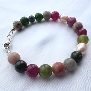 Könnte beinhalten: Ein Perlenarmband mit einer Vielzahl runder, polierter Edelsteine in Grün-, Rosa-, Grau- und Schwarztönen, durchsetzt mit zwei blassrosa Perlen. Das Armband wird mit einem silberfarbenen Karabinerverschluss und einem Biegering befestigt.
