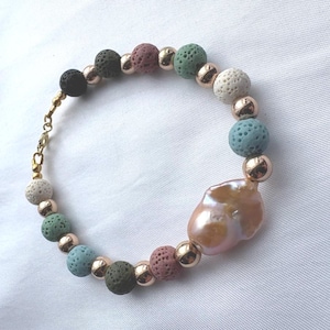 May include: A beaded bracelet featuring a large, irregular, iridescent peach-colored baroque pearl as the focal point. The band consists of alternating porous lava stone beads in shades of black, green, blue, and pink, interspersed with small gold-tone metal spheres and a gold-tone lobster claw clasp.