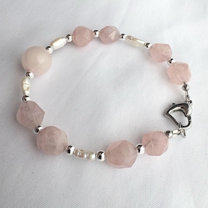 Könnte beinhalten: Ein Armband mit facettierten, blassrosa Rosenquarzperlen, kleinen weißen Süßwasserperlen und runden silberfarbenen Metallzwischenperlen. Das Schmuckstück wird mit einem herzförmigen Karabinerverschluss aus silberfarbenem Metall geschlossen.