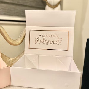 May include: White gift box with a gold foil accent and the text "Will you be my bridesmaid?" on the front. The box is open and has a white interior.