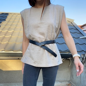 Pode incluir: Um top bege sem mangas com um cinto preto na cintura. O top tem um decote em V subtil e ombros almofadados. O cinto é de couro preto com uma fivela prateada. A pessoa está usando jeans escuros e botas pretas.